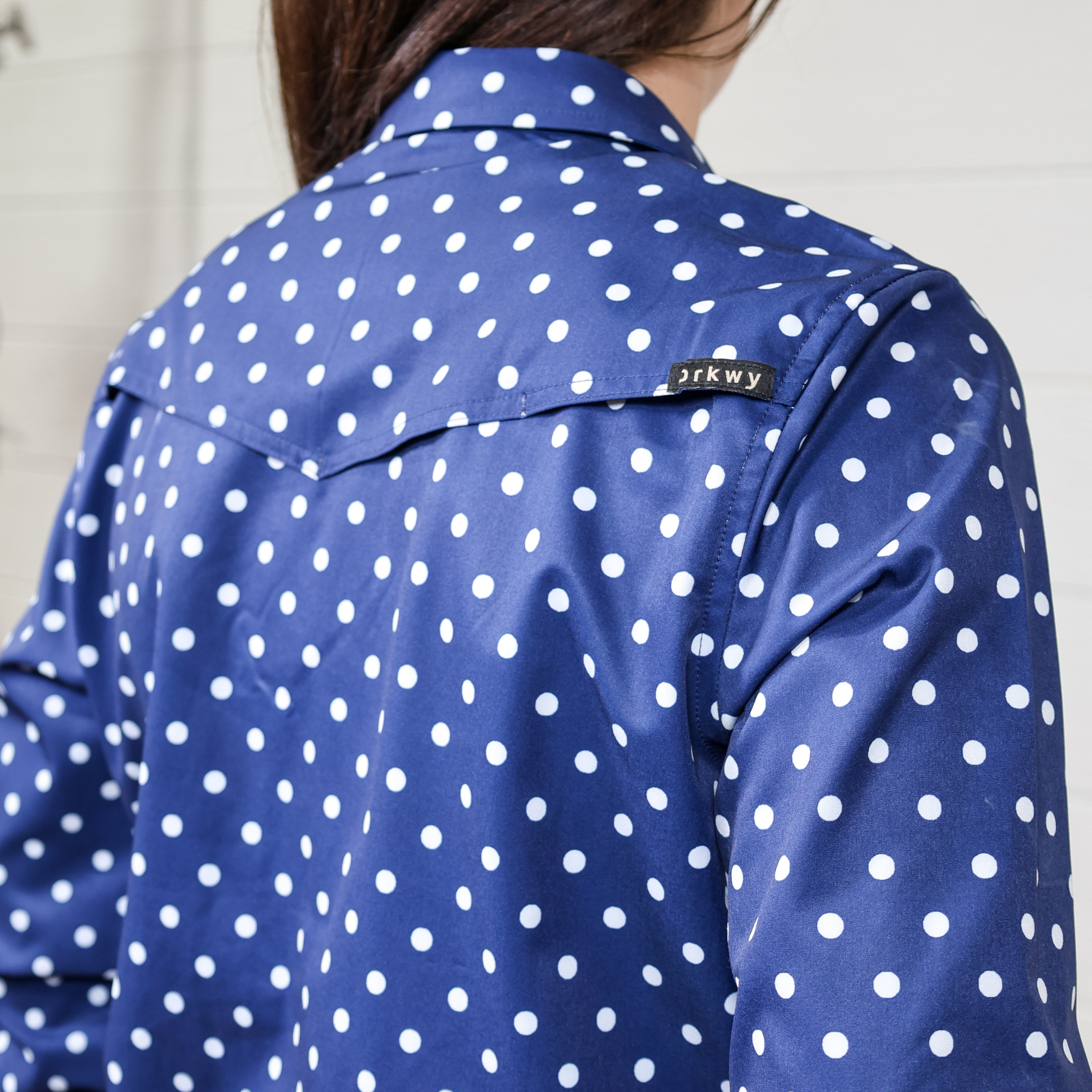 Blue polka dot shirt with a brand label on the back against a white background. back facing.