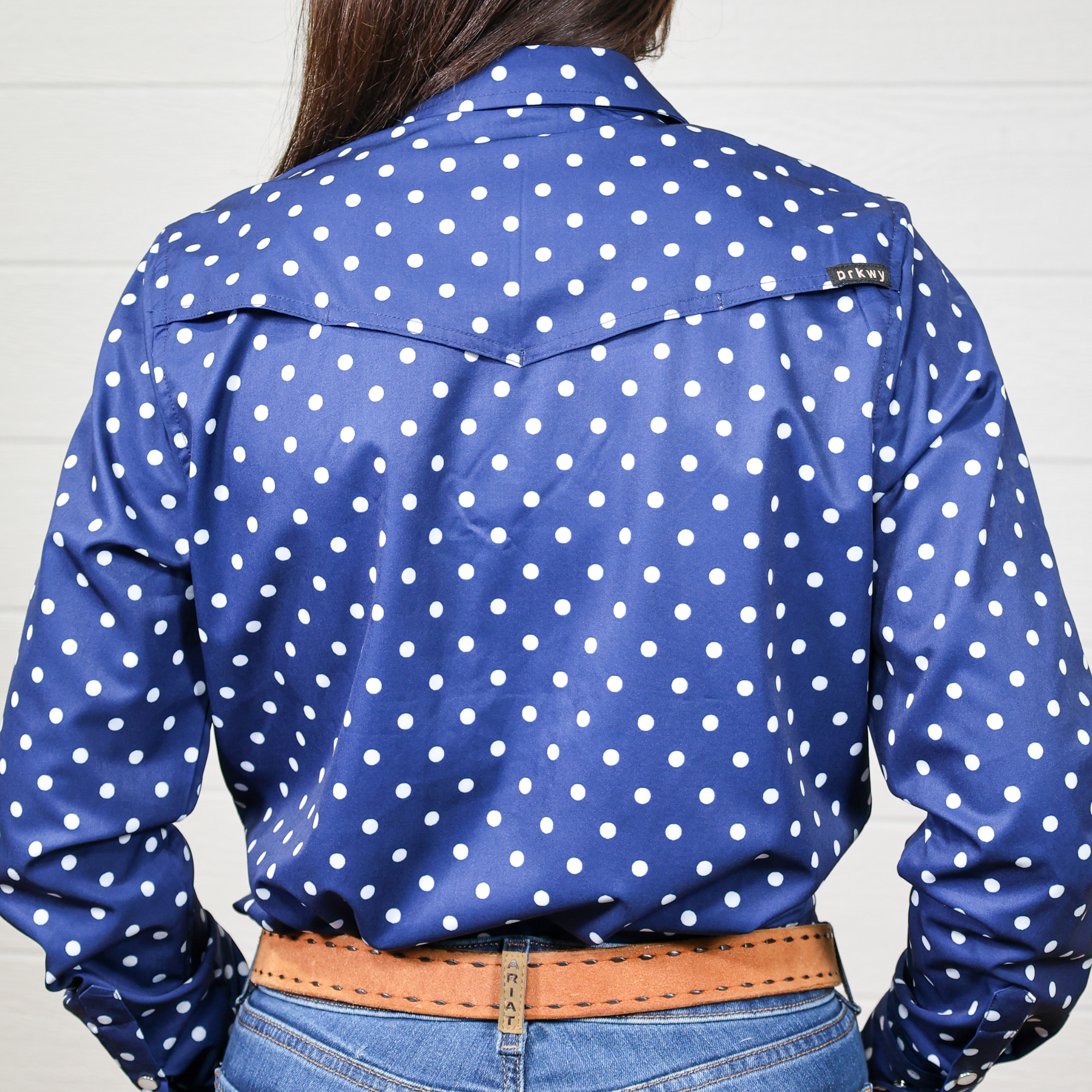 Blue polka dot shirt worn by a person on a white background. back facing.