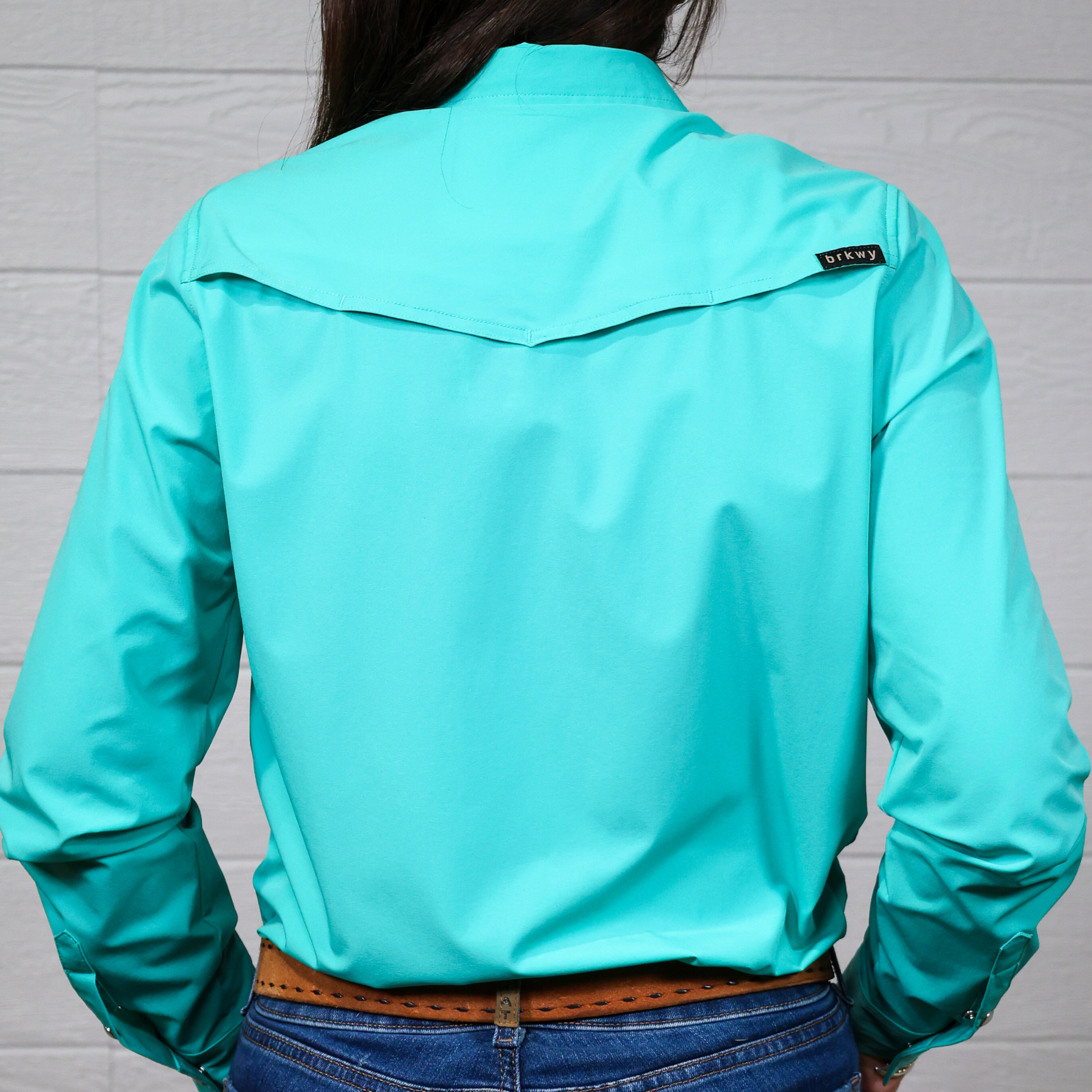 Person wearing a turquoise button down with a brand logo on the back against a white background. back facing.