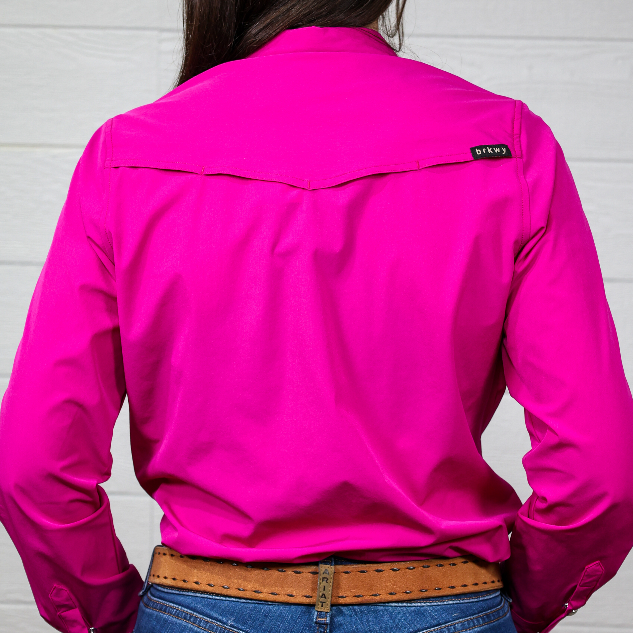 Person wearing a bright pink shirt with a brand logo on the back, standing against a white wall. back facing,