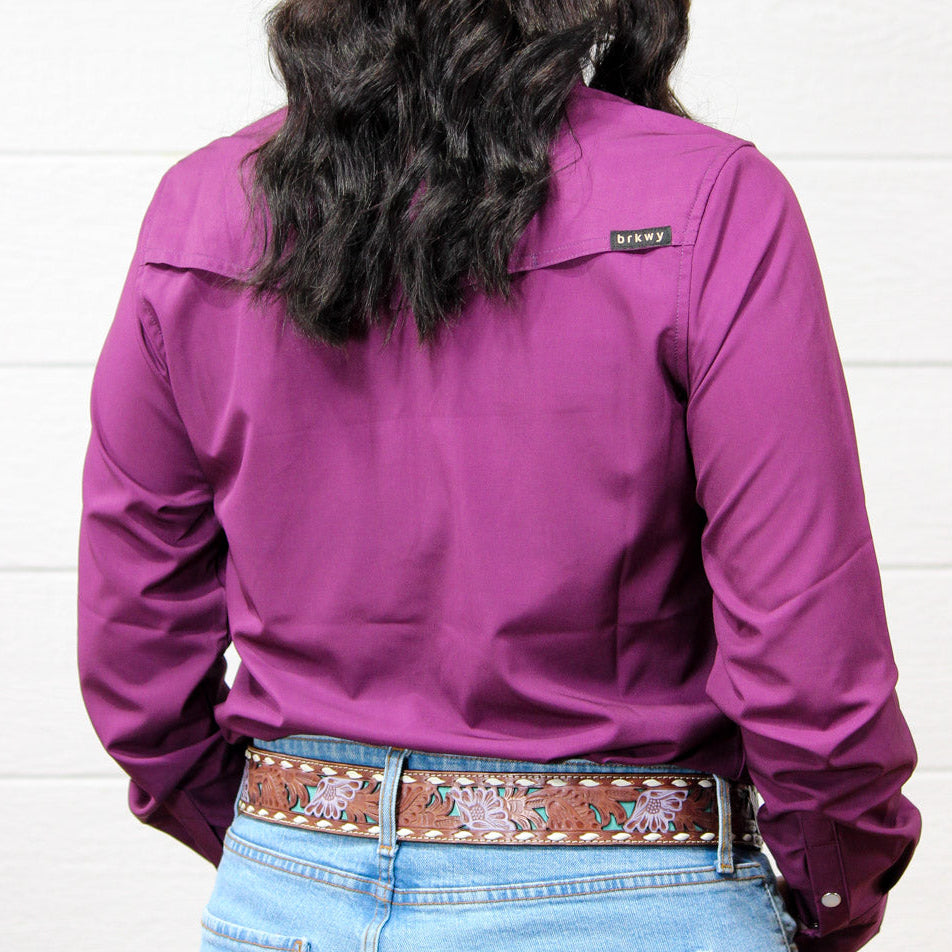 A person shown from the back wearing a long-sleeve magenta-purple button-down shirt, light-wash blue jeans, and a brown embroidered belt against a white wood-paneled wall.