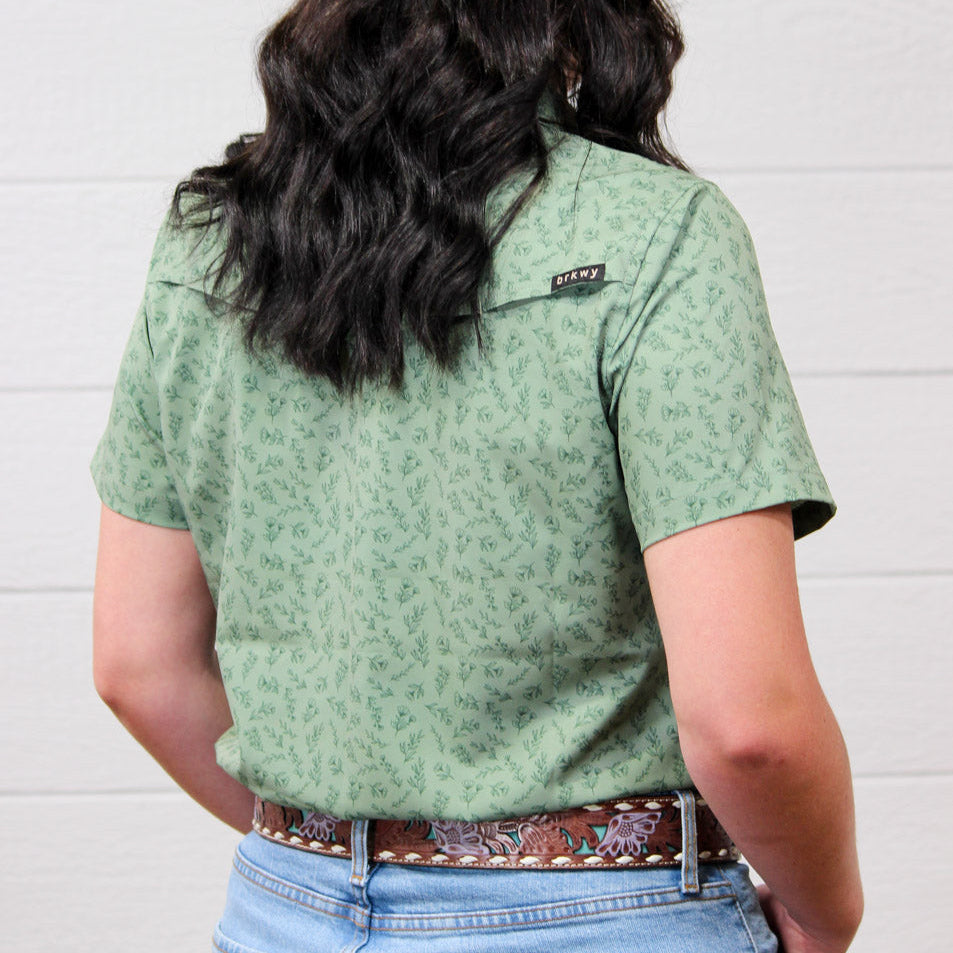 A person shown from the back wearing a sage green short-sleeve button-up shirt with a small allover floral pattern, paired with a brown patterned belt and blue jeans against a white wood-paneled wall.