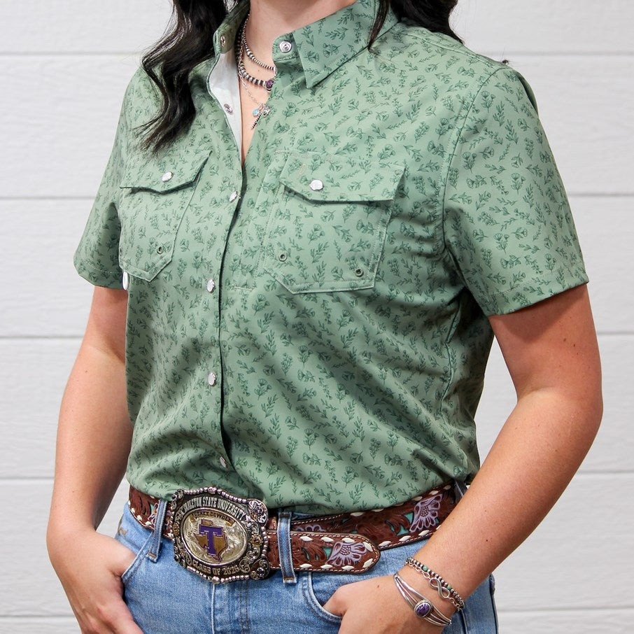 A woman wearing a sage green short-sleeve western-style button-up shirt with a small floral print, paired with blue jeans and a brown western belt, standing against a white wood-paneled wall.