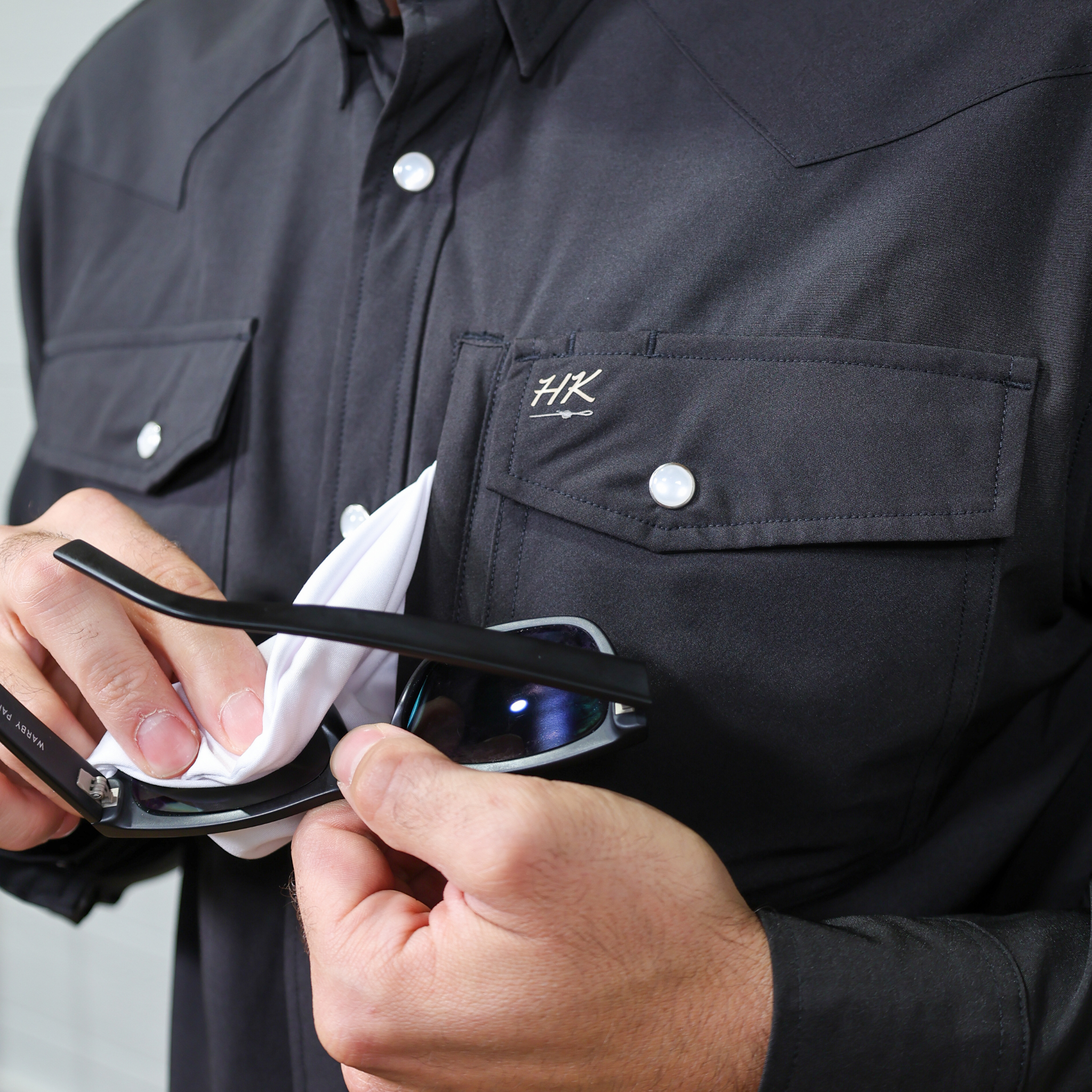 Person cleaning sunglasses with a cloth, attached to a  black shirt with a logo. 