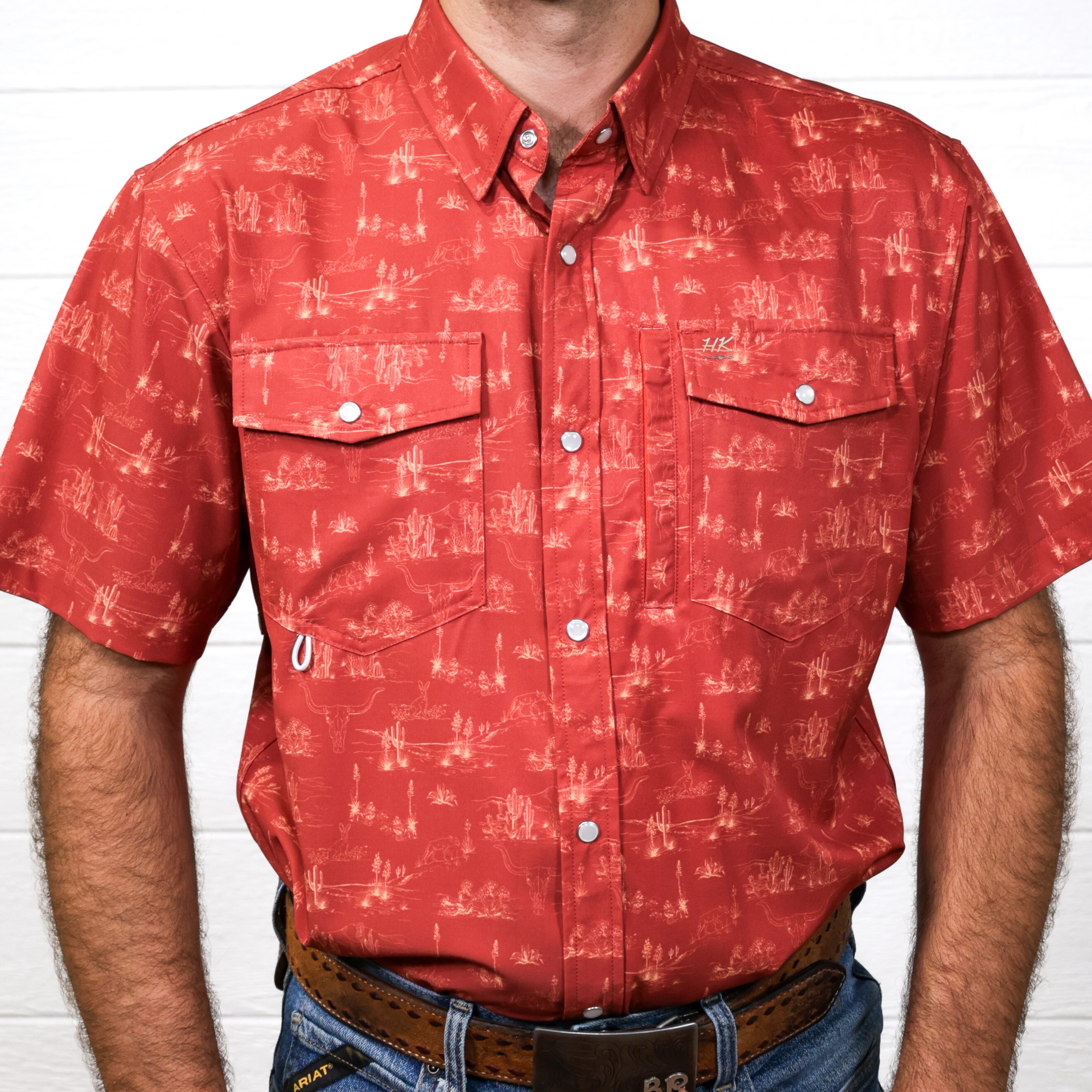 An orange-patterned short-sleeve shirt worn by a person against a white brick wall, showing the front.