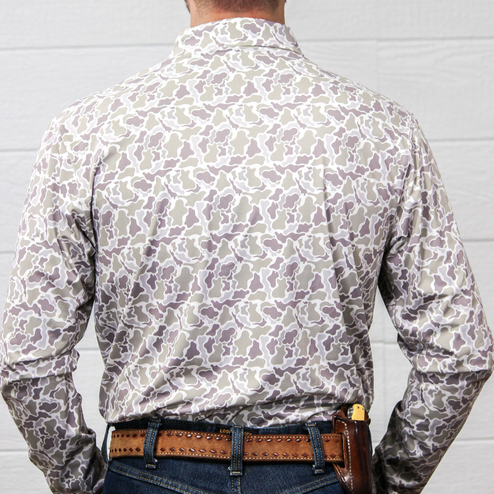 Back view of a man wearing a long-sleeve button-up shirt with a gray and white abstract, camo pattern, tucked into blue jeans with a brown studded belt.
