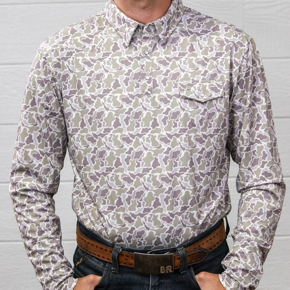 A long-sleeve button-down shirt with a beige, gray, and brown camouflage pattern, paired with a brown leather belt and dark jeans, standing against a white paneled wall.