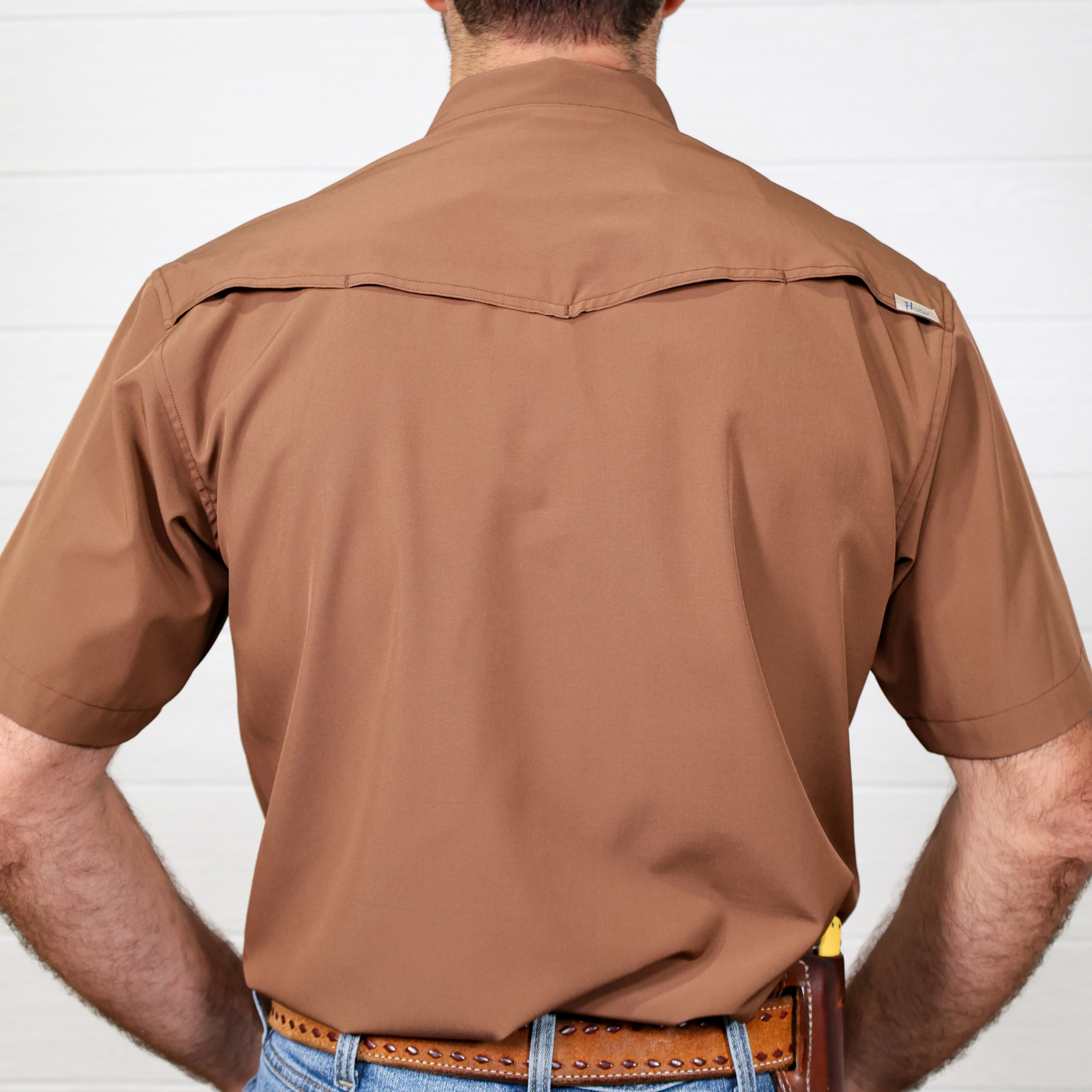 Man wearing a short sleeve brown shirt with a visible brand logo on a white background showing the back 