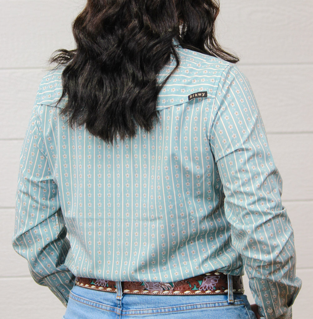 A person shown from the back wearing a light blue long-sleeve button-up shirt with a vertical geometric pattern in white and peach, paired with blue jeans and a patterned belt against a white paneled wall.