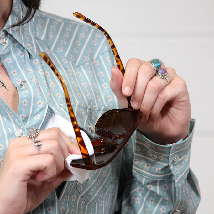 A person wearing a light blue and white patterned button-up shirt holds a pair of tortoiseshell sunglasses with dark lenses, using a white cloth to wipe the lenses. The person is wearing several rings.