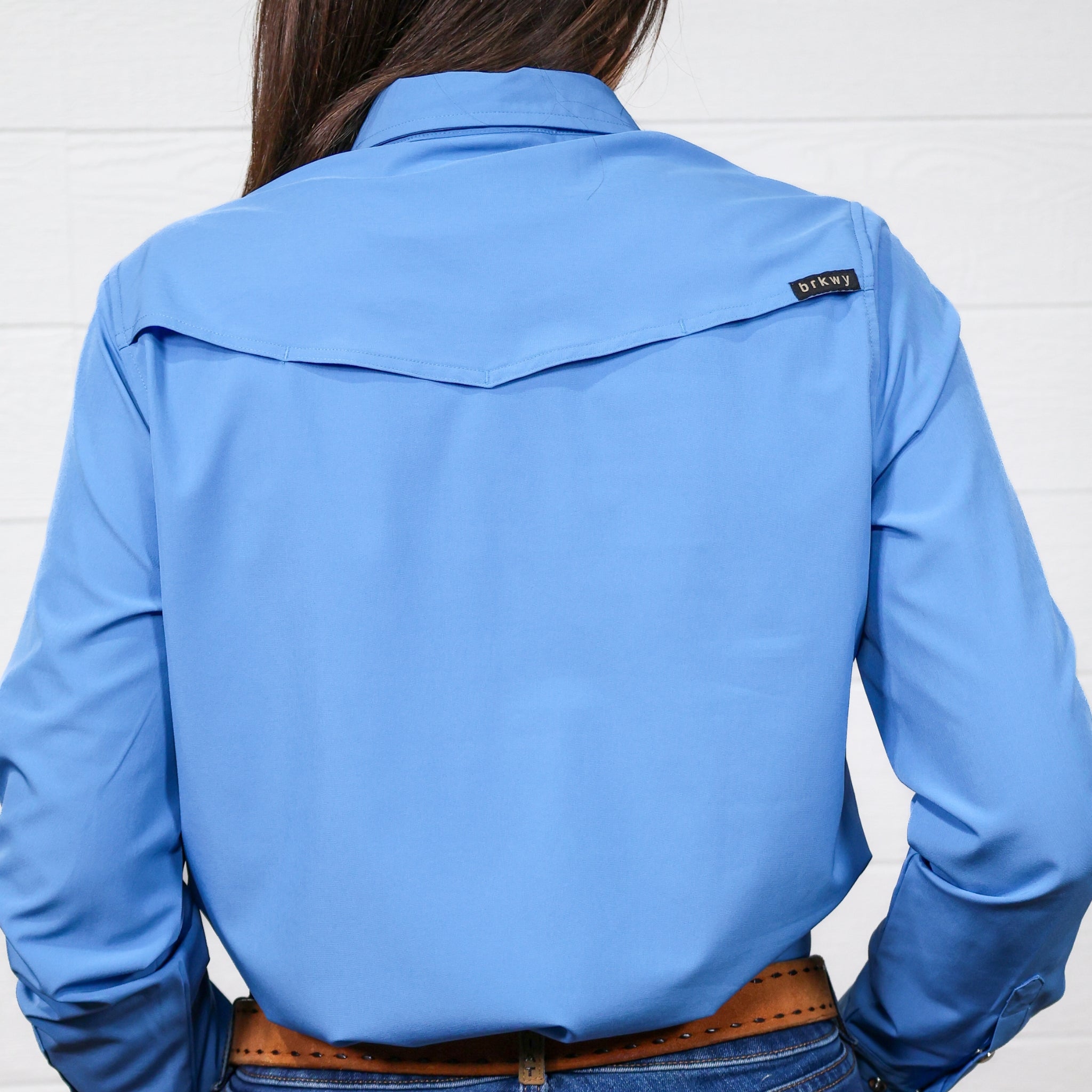 Back of Women wearing a denim blue pearl snap long sleeve top tucked in to blue jeans with a belt in front of a white wall. 