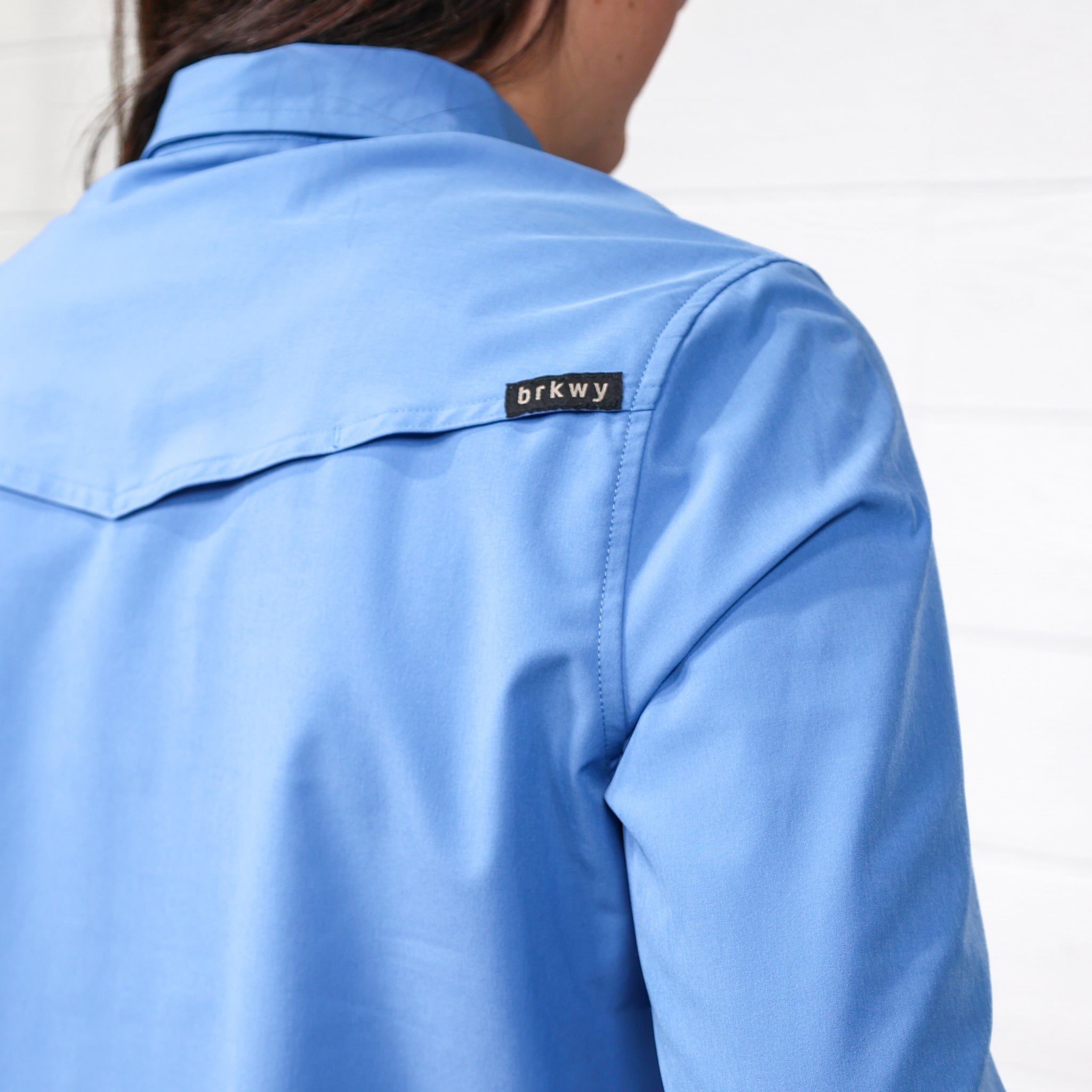 Detail Of Women wearing a denim blue pearl snap long sleeve top tucked in to blue jeans with a belt in front of a white wall. 