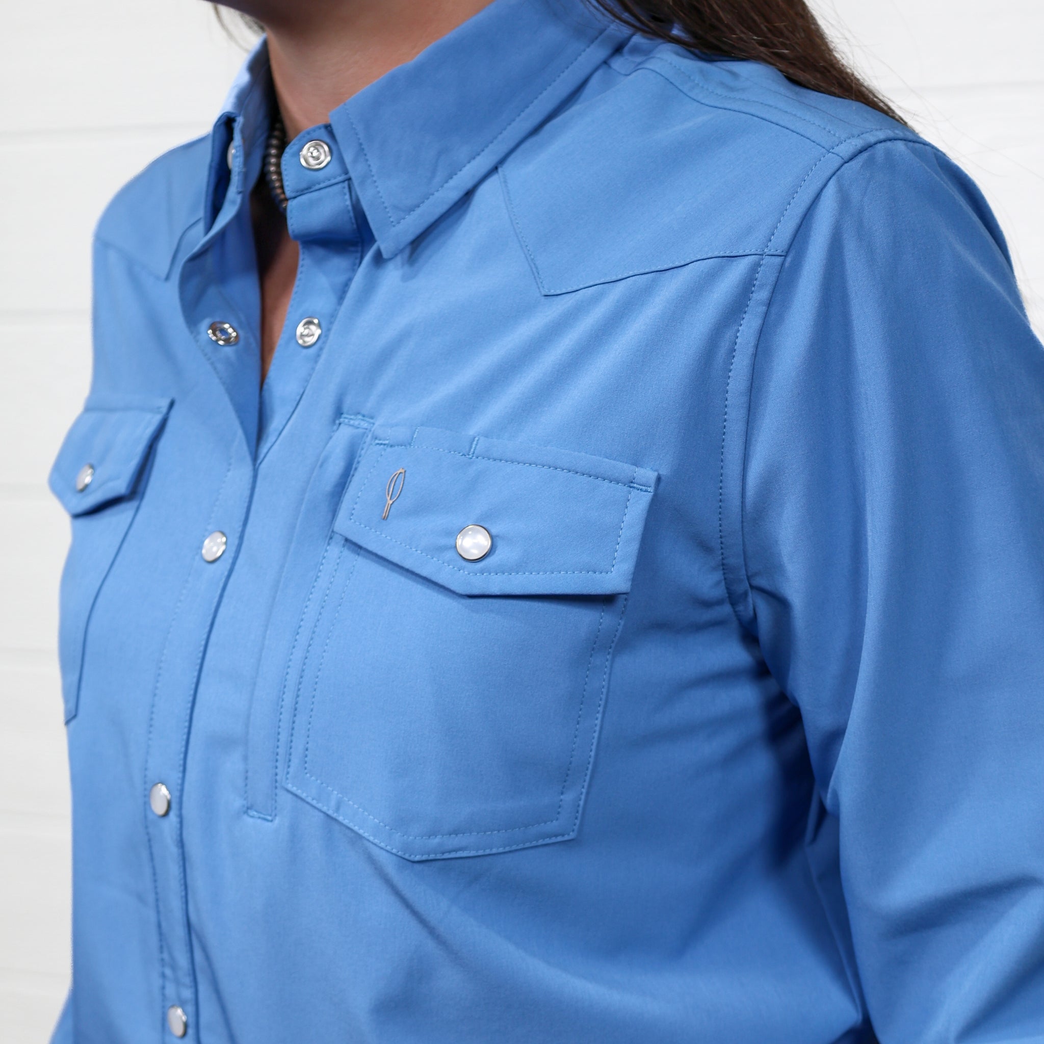 Detail of Women wearing a denim blue pearl snap long sleeve top tucked in to blue jeans with a belt in front of a white wall. 