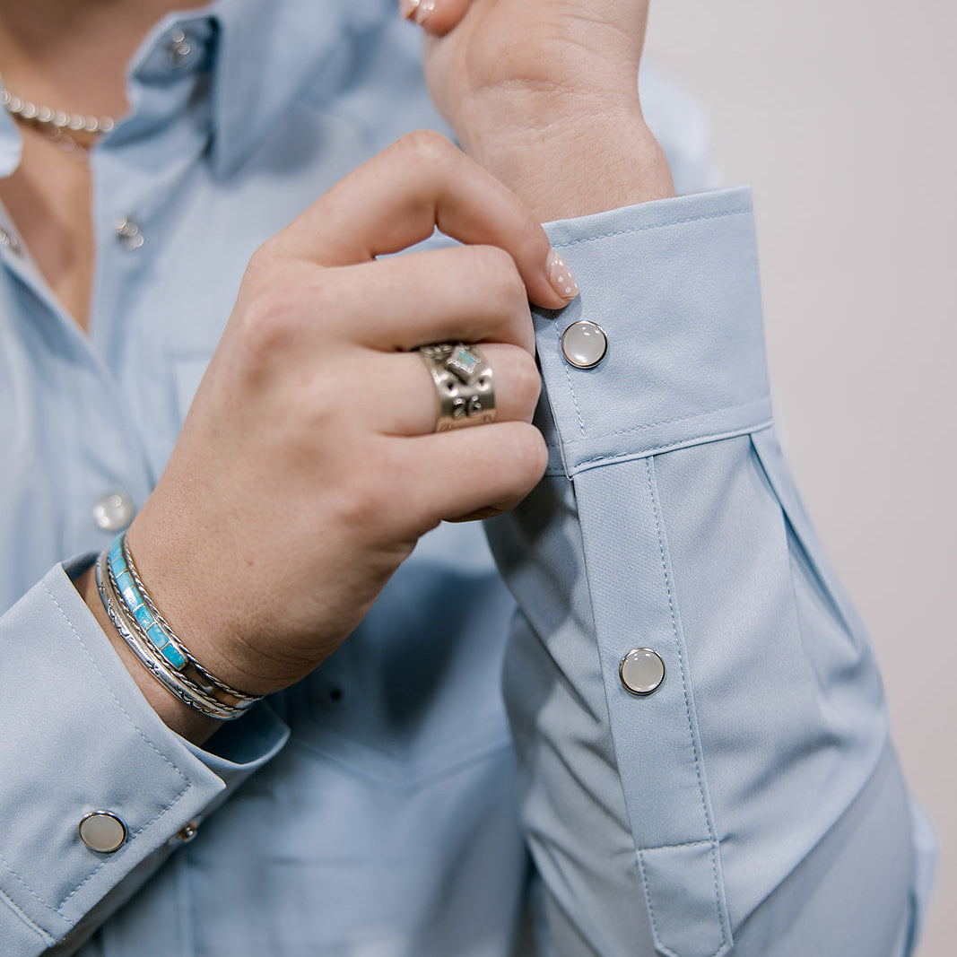 Person buttoning a light blue shirt with a neutral background showing the buttons 