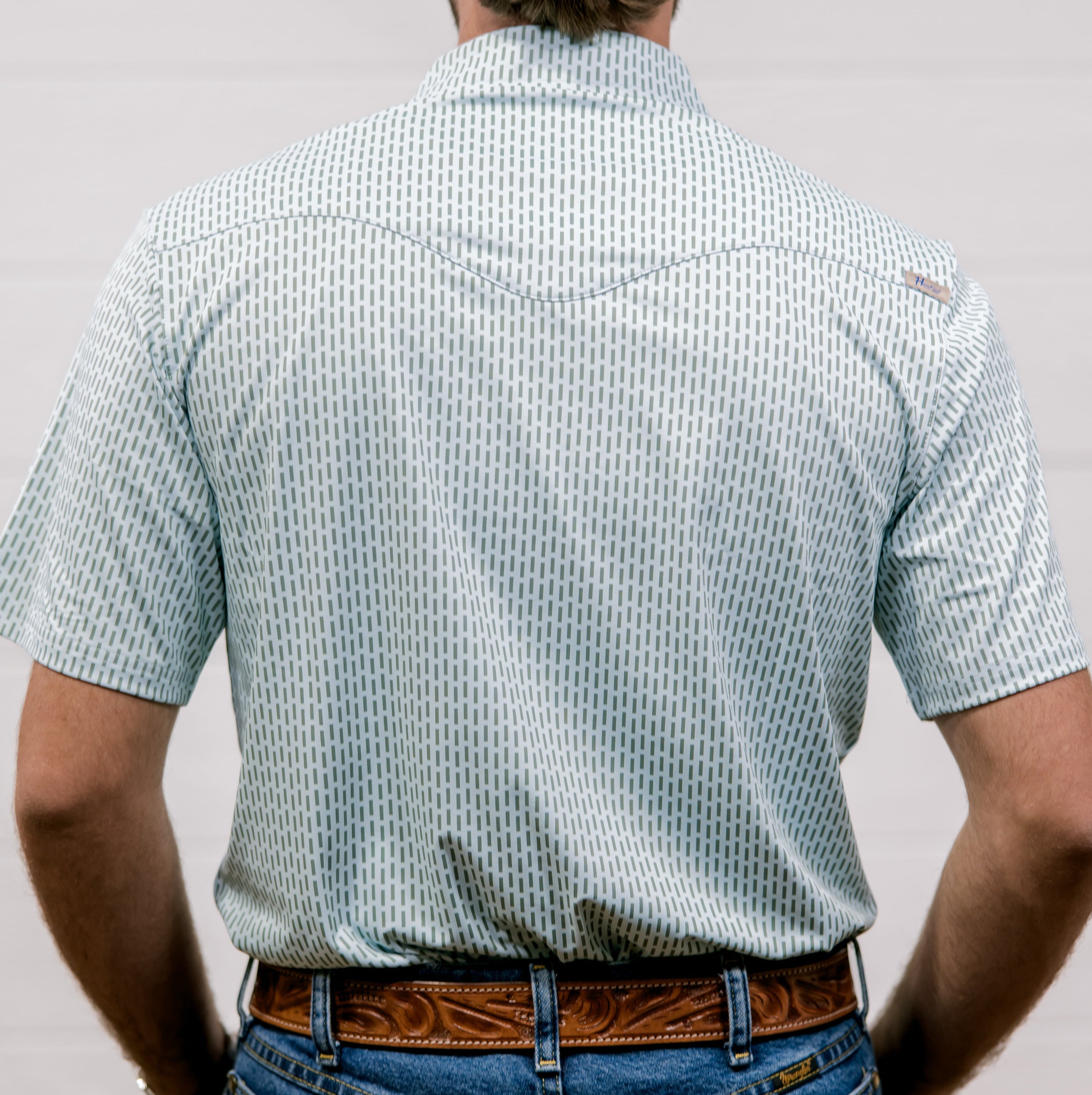 Man wearing a light blue and green  patterned shirt and blue jeans against a plain background. back facing.