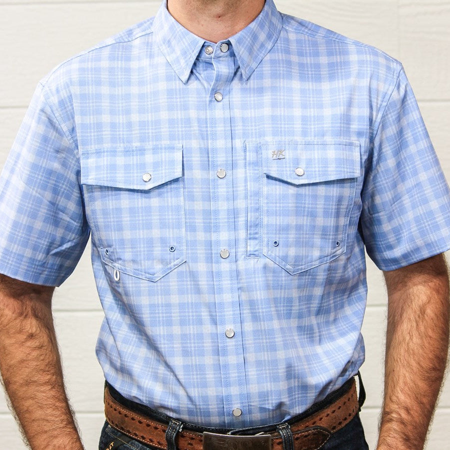 A man wearing a light blue and white plaid short-sleeve button-down shirt with two chest pockets, paired with a brown leather belt and dark jeans, standing against a white wood-paneled wall.