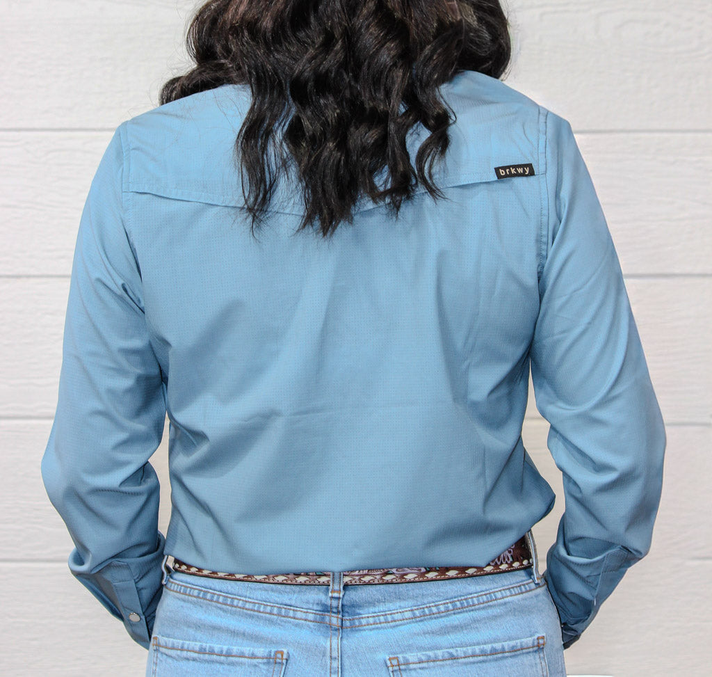 A person shown from the back wearing a light blue long-sleeve button-up shirt and light-wash jeans, standing against a white wood plank wall. A small black brand tag reading 'STANLEY' is visible near the upper right shoulder.