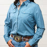 A woman wearing a light blue long-sleeve western-style button-up shirt with two chest pockets, paired with blue jeans and a brown patterned belt with a large decorative buckle, standing against a white wood-paneled wall.