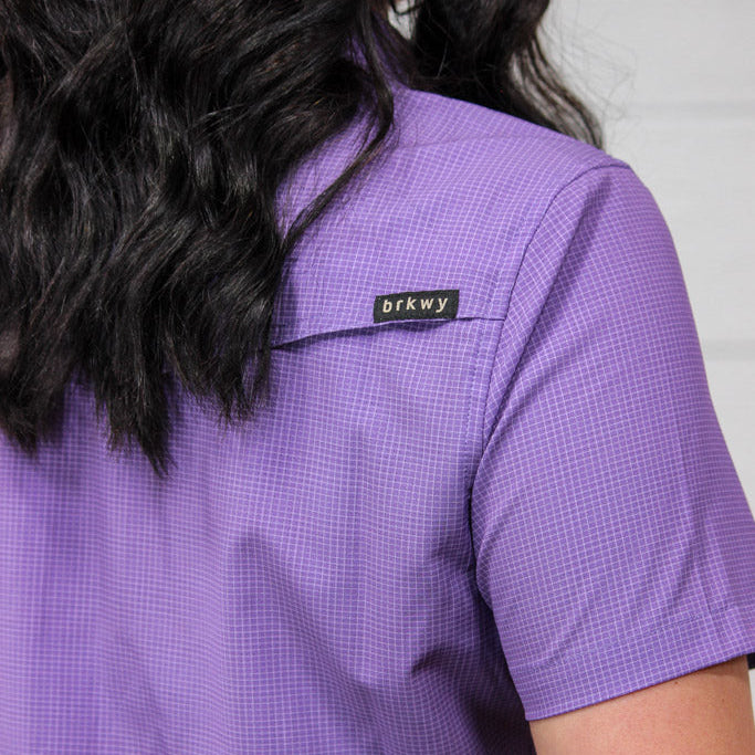 Close-up back view of a person wearing a short-sleeve purple gingham scrub top with a small black 'brkwy' brand tab on the upper back, against a light neutral background.