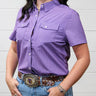 A woman wearing a short-sleeve purple button-up shirt with two chest pockets, paired with blue jeans and a western belt, standing against a white paneled wall.