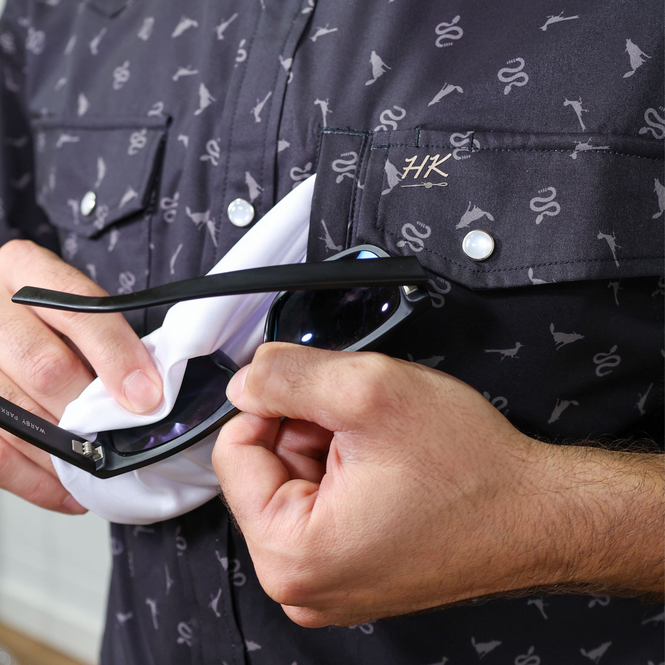 Person cleaning sunglasses with the lens cloth, wearing a dark shirt with a pattern.