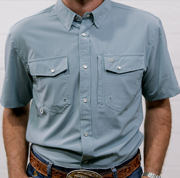 Man wearing a light blue rangevent shirt and jeans with a belt and watch, standing against a plain background