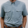 Man wearing a light blue rangevent shirt and jeans with a belt and watch, standing against a plain background