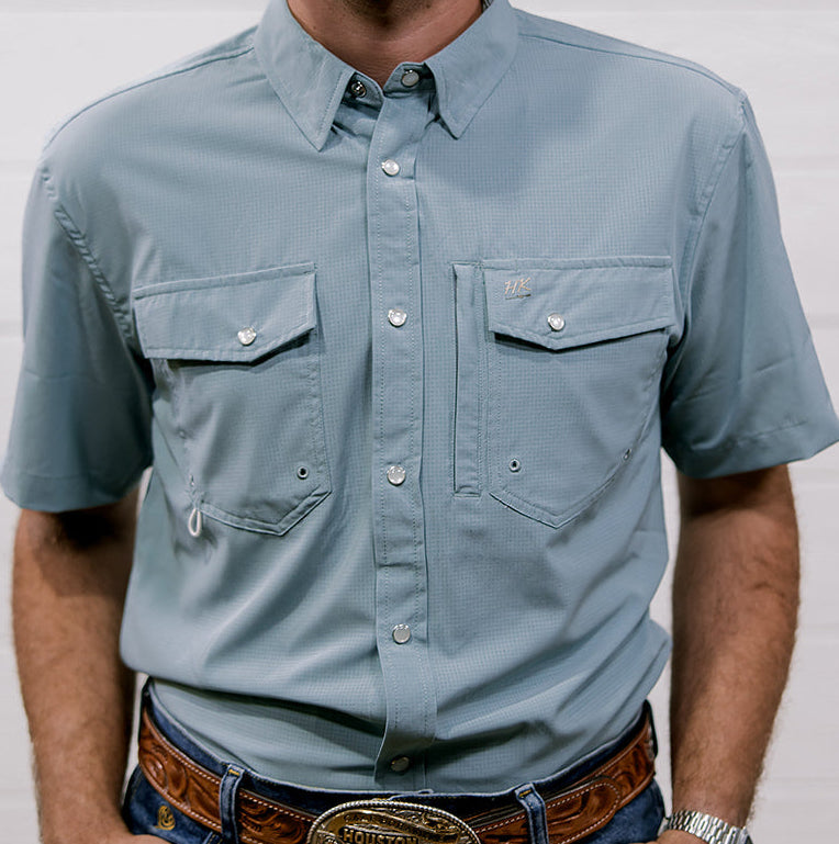 Man wearing a light blue rangevent shirt and jeans with a belt and watch, standing against a plain background