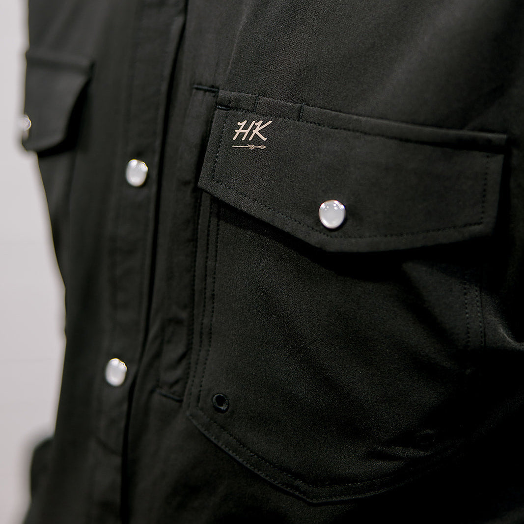 Close-up of a black shirt with a brand logo on a plain background. Pocket detail.