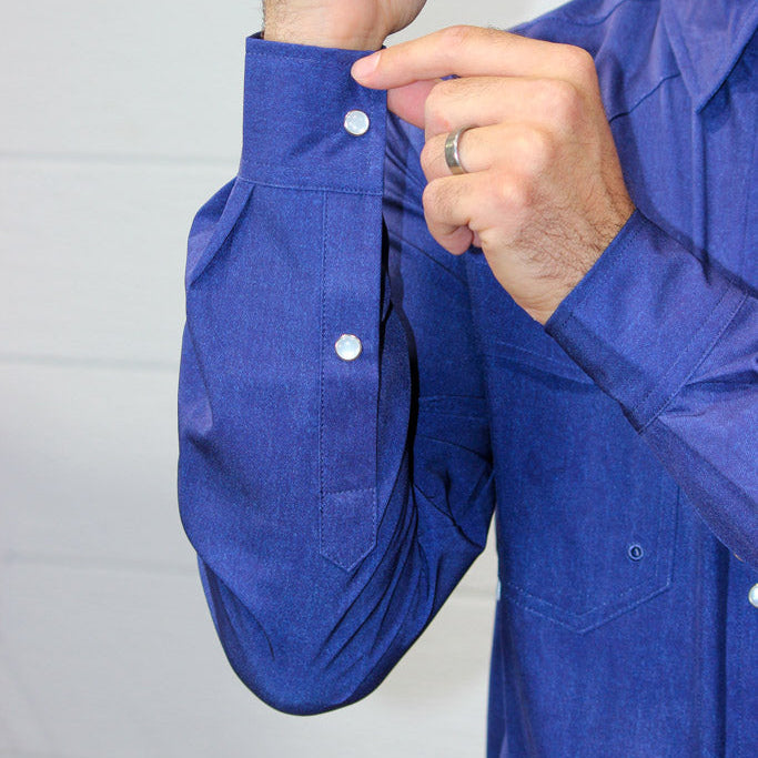 A man wearing a blue long-sleeve button-down shirt, demonstrating how to fold the cuff and button it, against a white paneled wall.