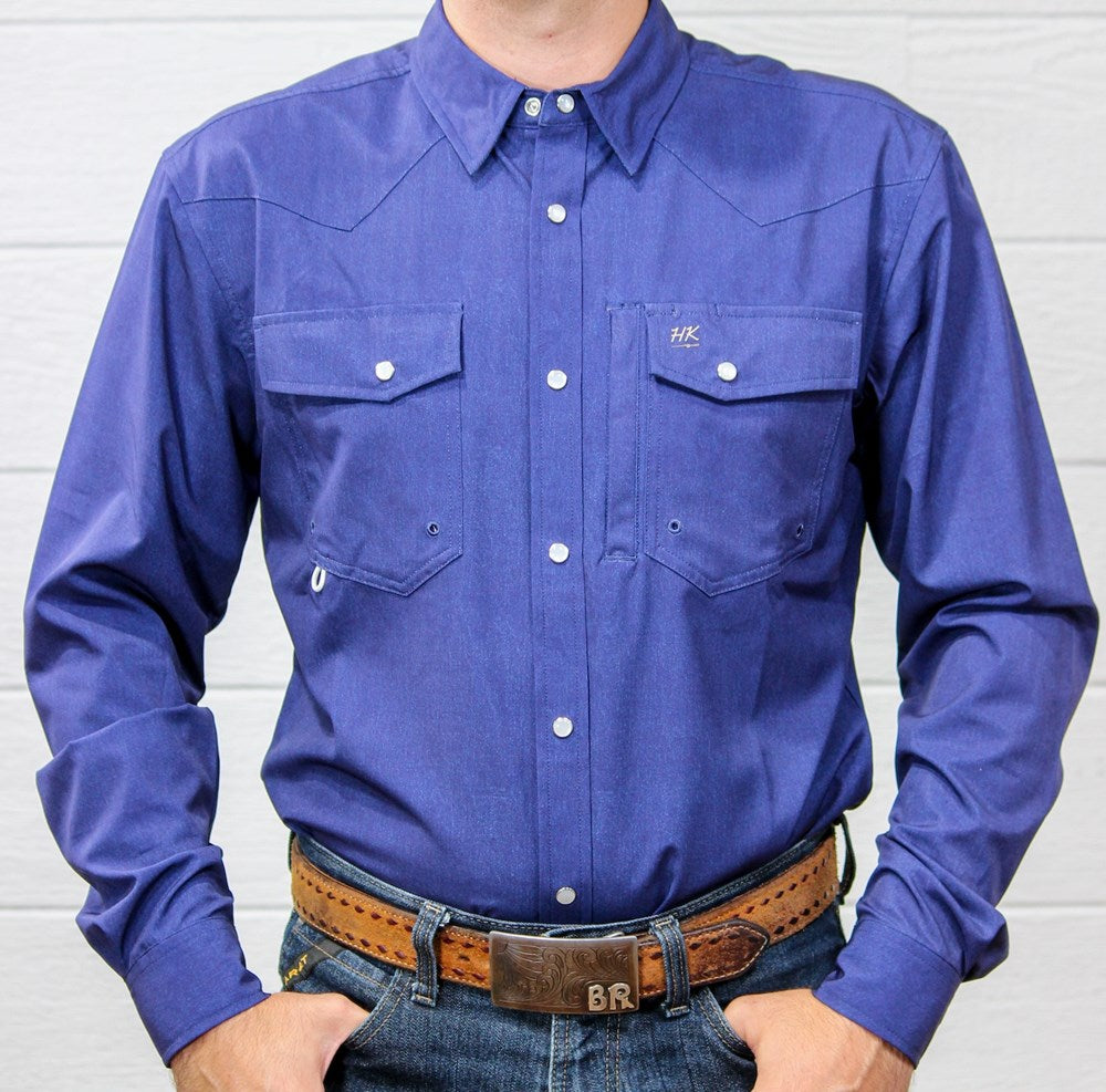 A man wearing a long-sleeve western-style button-down shirt in a medium periwinkle blue color, paired with a brown leather belt and blue jeans, standing against a white wood-paneled wall.