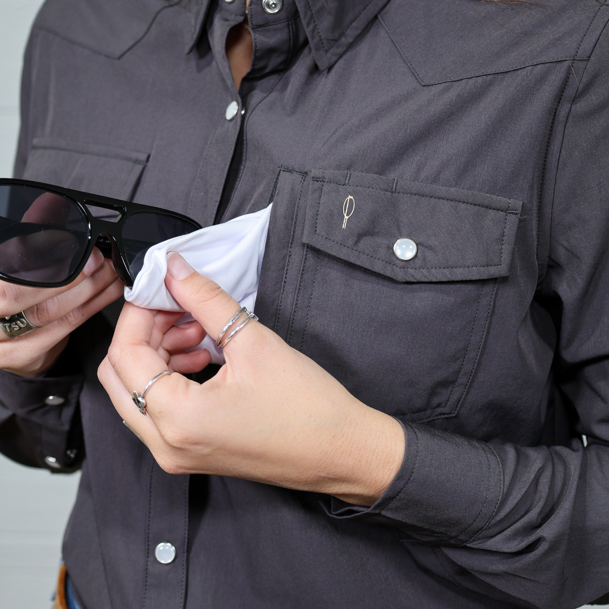 Person wearing a dark gray  women's button down with a pocket, holding sunglasses and a white cloth attached to the shirt. Foward facing