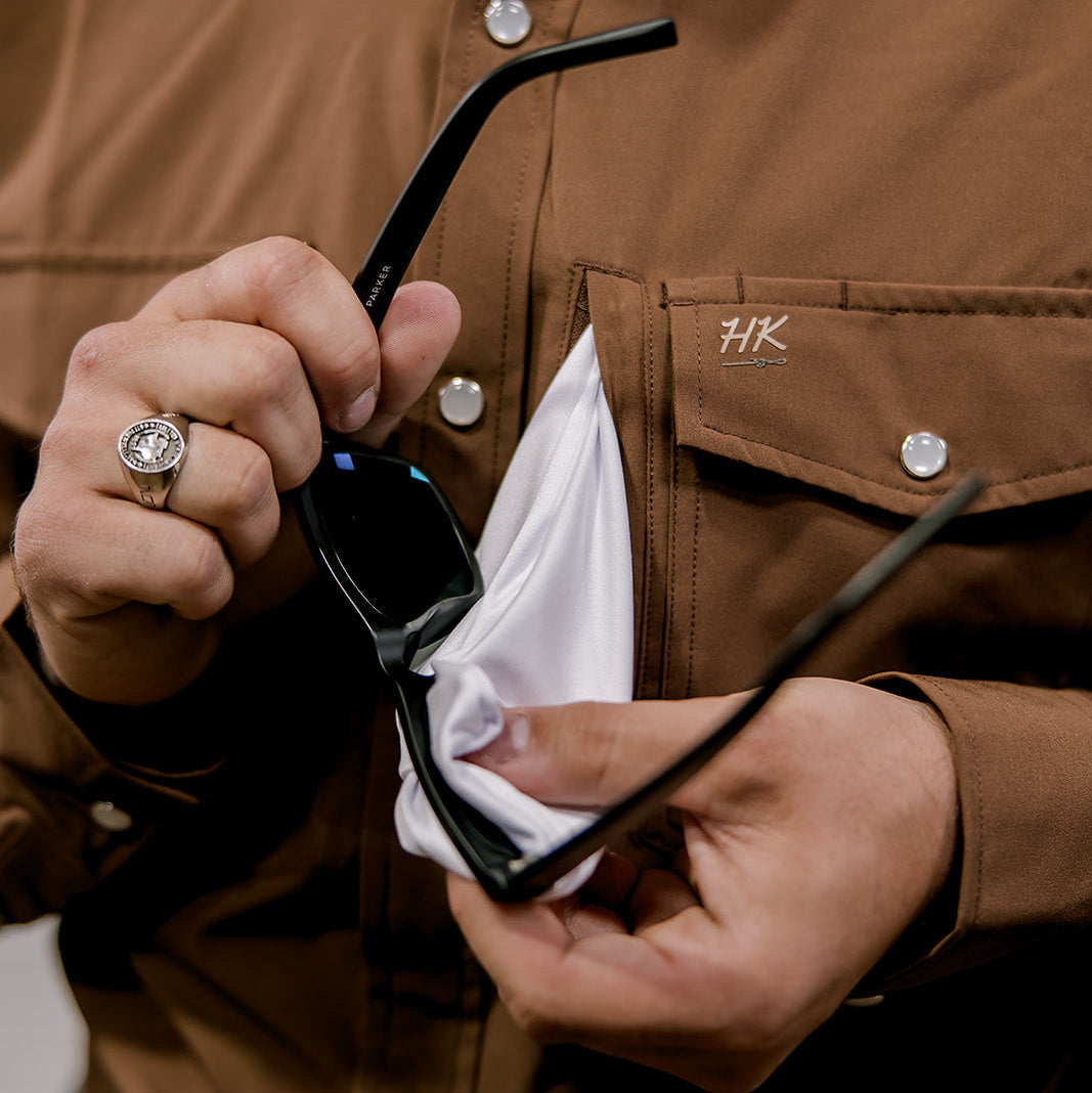 Person cleaning sunglasses with a cloth, wearing a brown shirt with a logo.