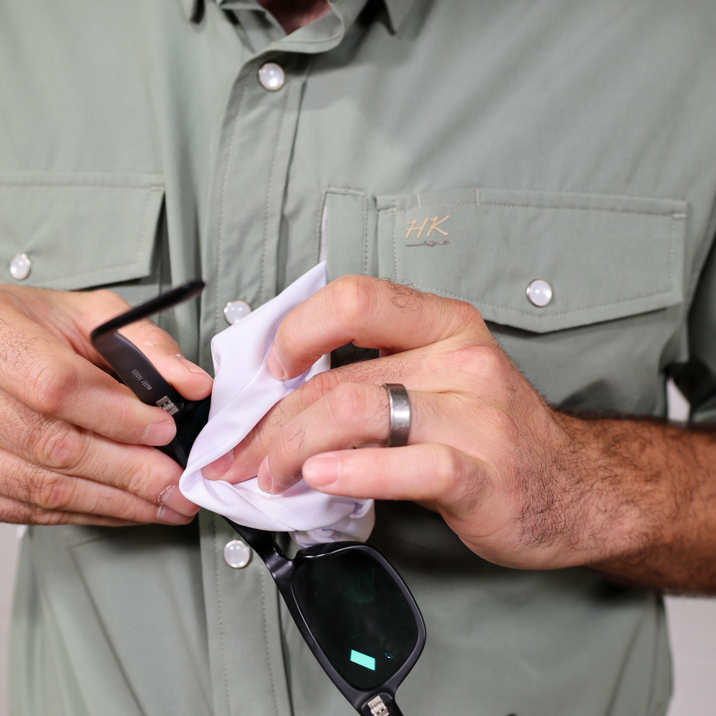 Person cleaning sunglasses with lens cleaning cloth, wearing a green shirt with a brand logo.