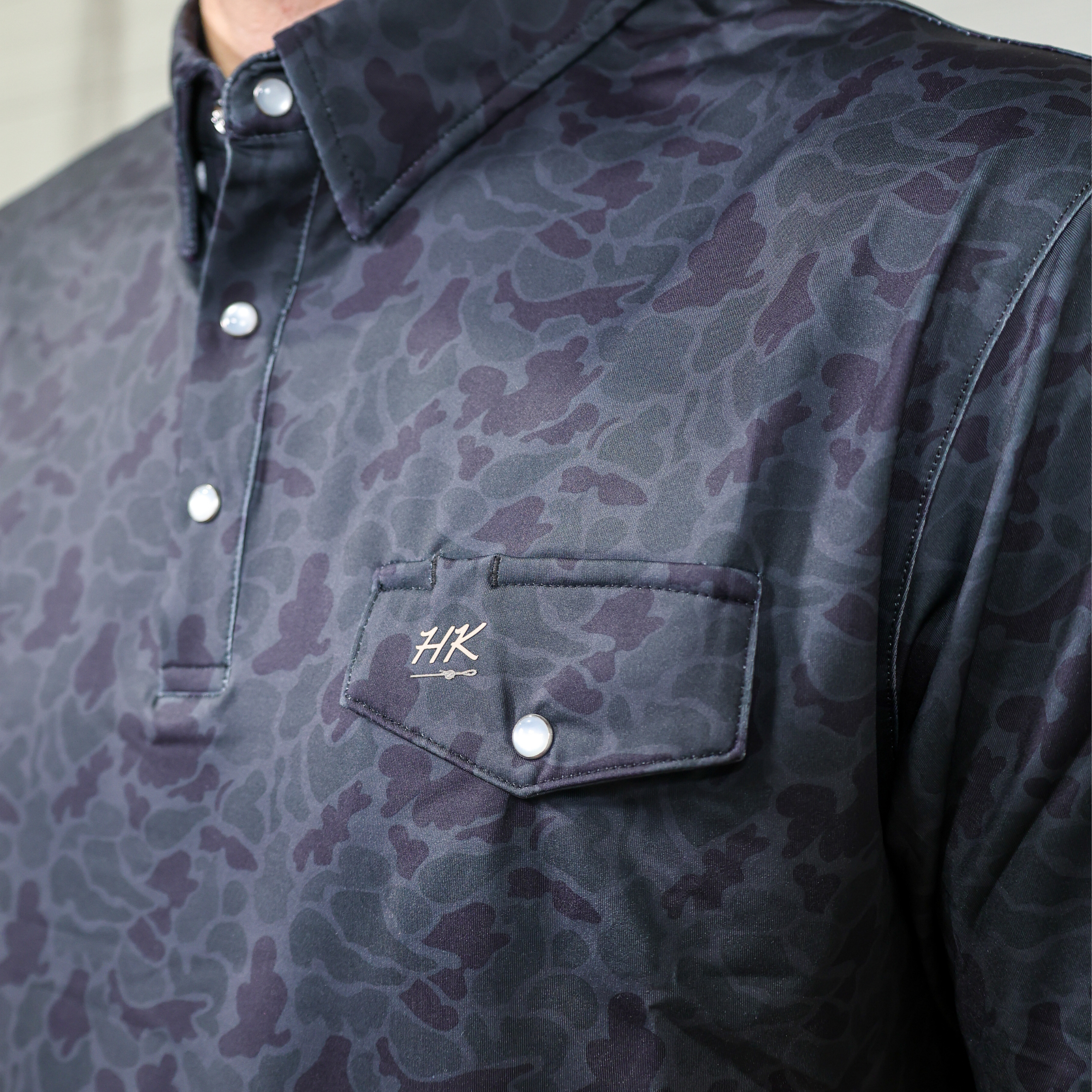 Dark camo patterned polo with a brand logo on the chest pocket against a neutral background