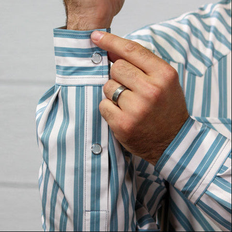 A close-up of a man's hands adjusting the cuff of a blue and white striped long-sleeve button-down shirt against a light gray paneled wall.