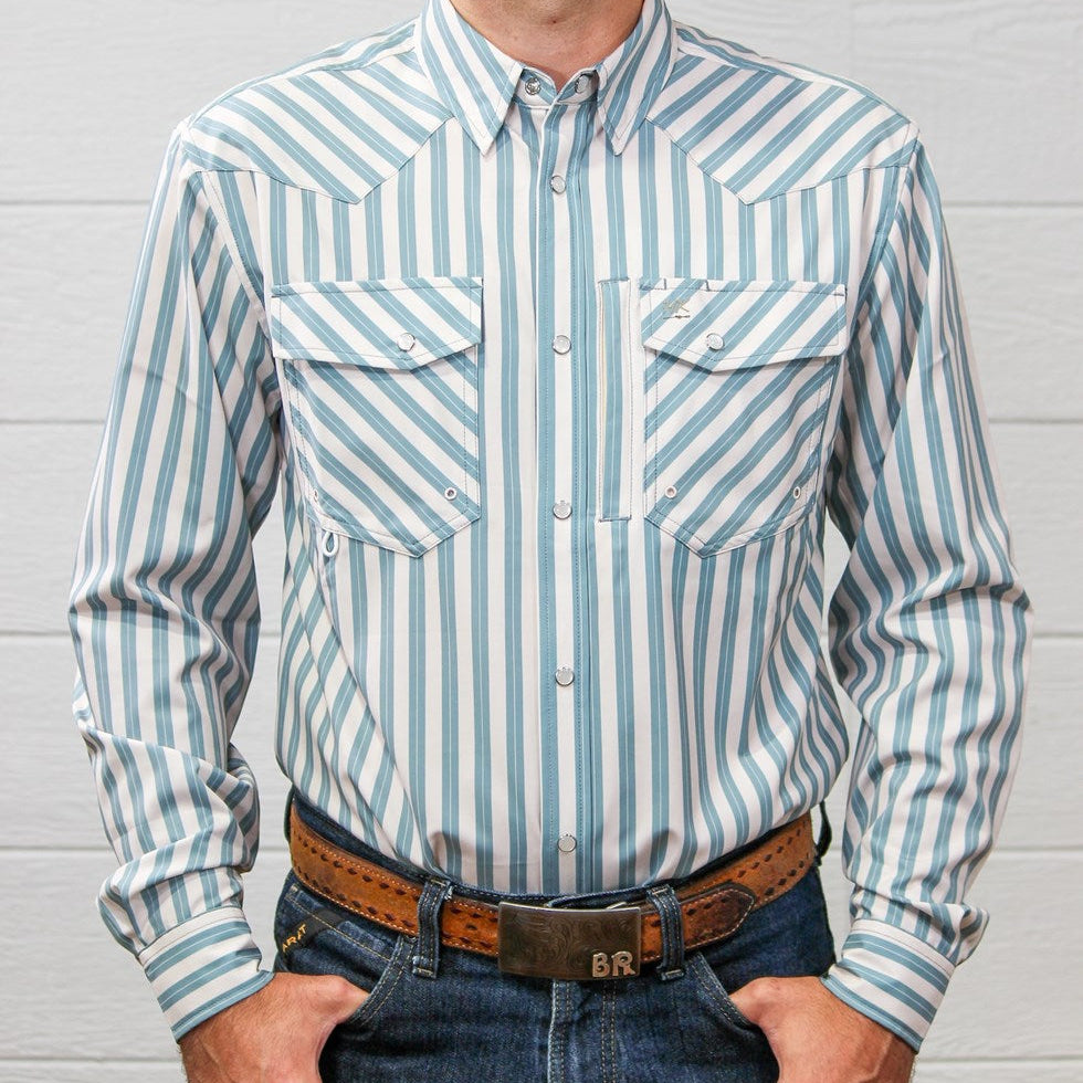 A man wearing a light blue and white vertically striped western long-sleeve shirt, paired with blue jeans and a brown leather belt, standing against a white wood-paneled wall.