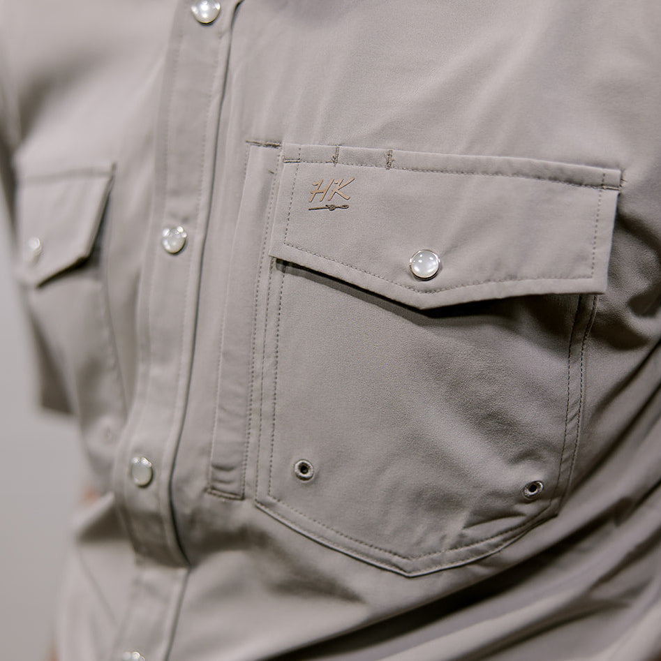 Close-up of a gray button-up shirt with a brand logo on a plain background. Side Facing 
