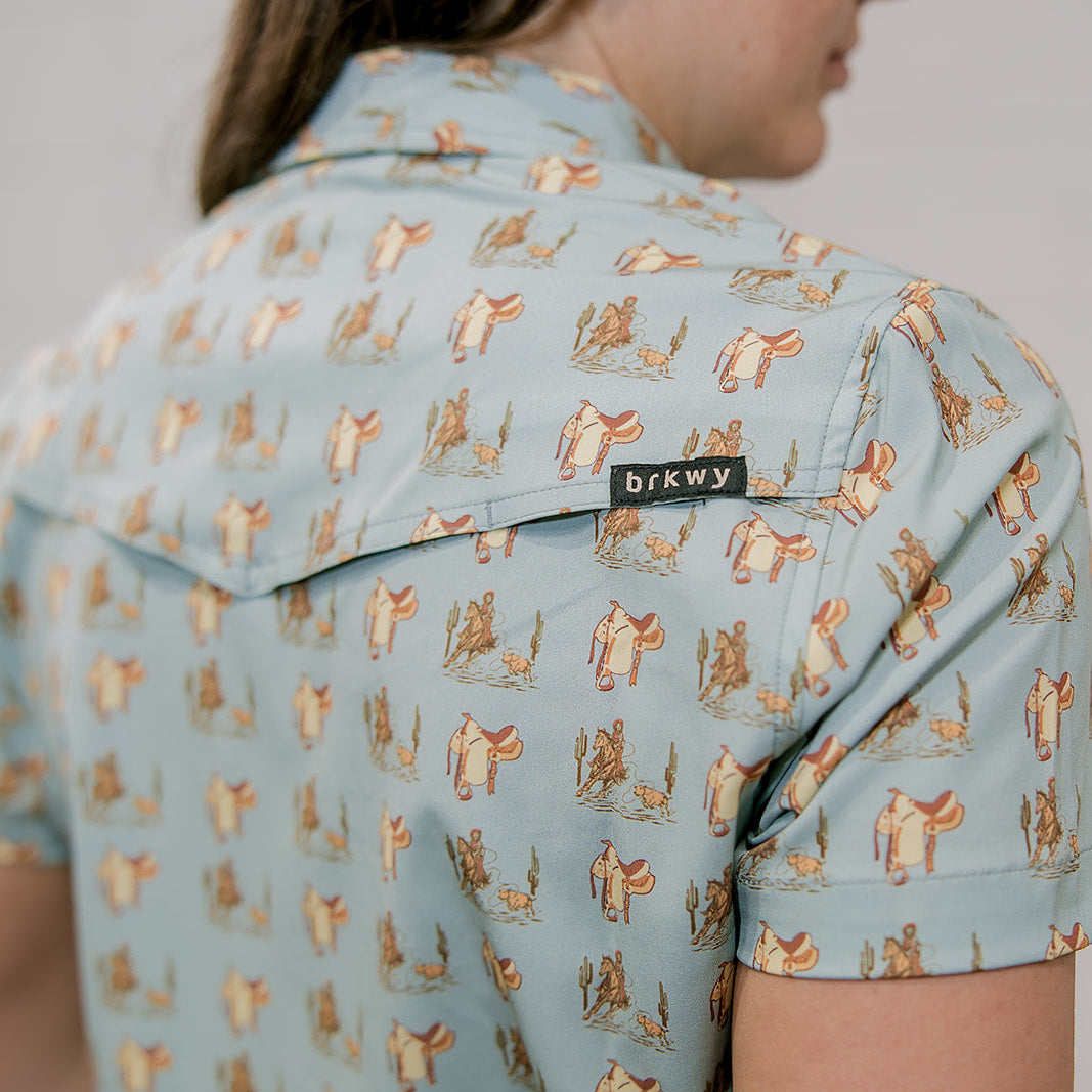 Person wearing a light blue shirt with a western pattern and 'brkwy' brand a label and vent .
