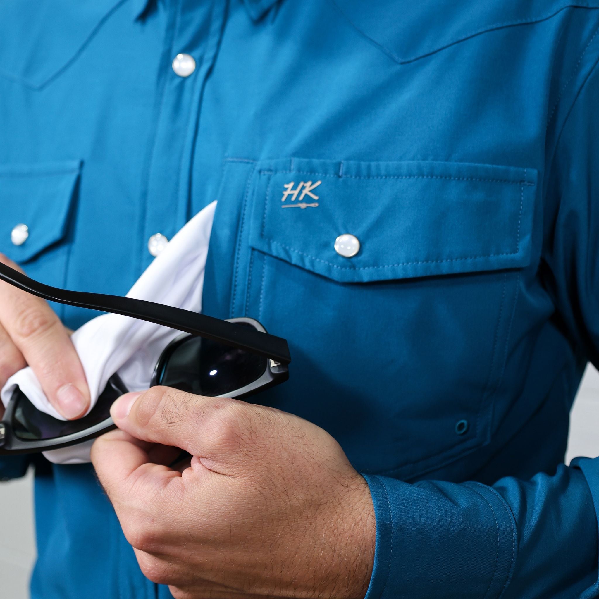 Person wearing a blue shirt with HK logo holding sunglasses showing the lens cloth 