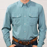 A man wearing a light blue-green long-sleeve western button-down shirt with two chest pockets, paired with a brown belt and dark jeans, standing against a white wood-paneled wall.