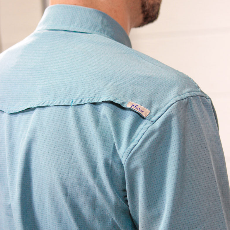 Close-up back view of a man wearing a light blue textured long-sleeve button-down shirt, showing the collar, shoulder, and a small visible Columbia brand tag on the upper shoulder.