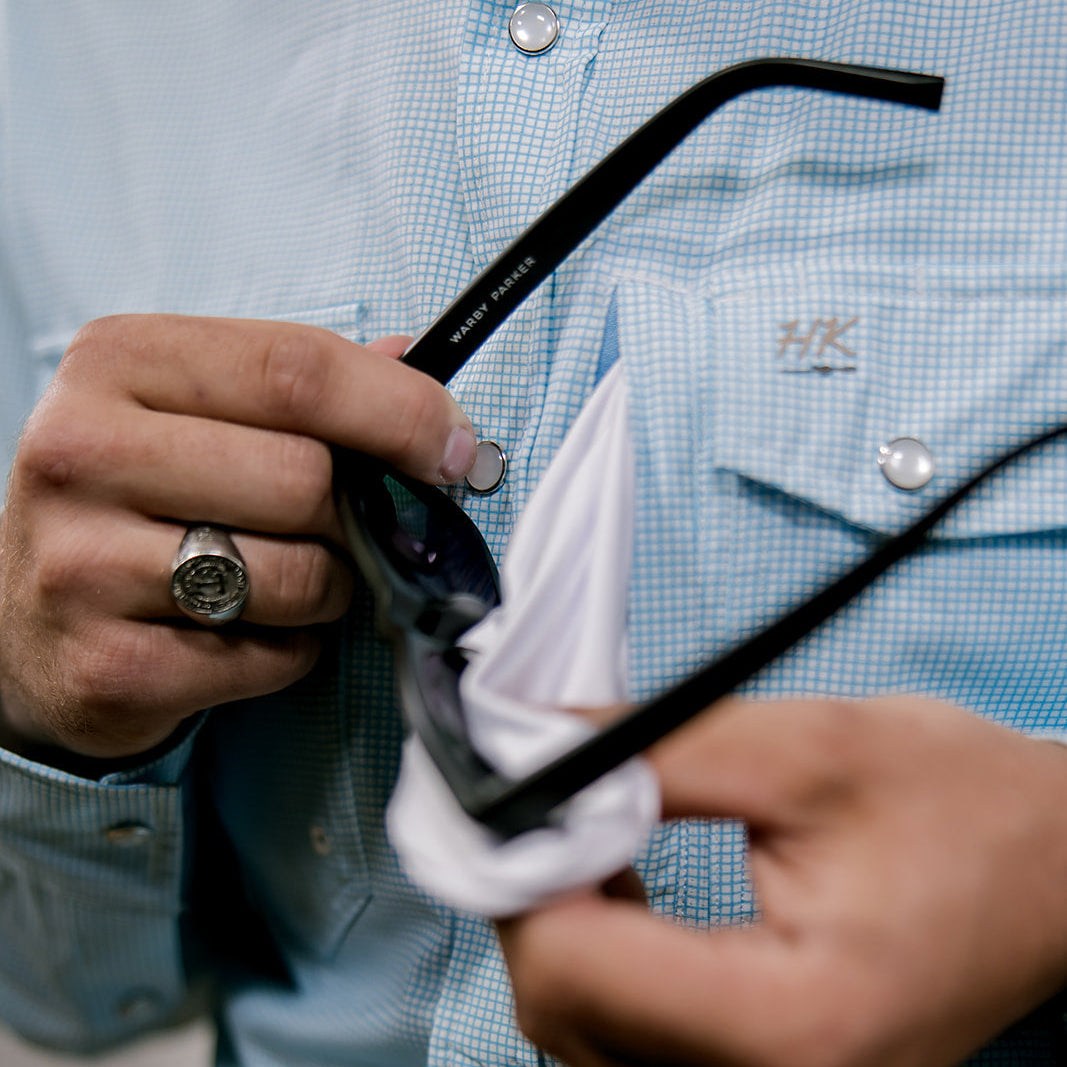 Person cleaning sunglasses with a white cloth, wearing a blue checkered shirt.