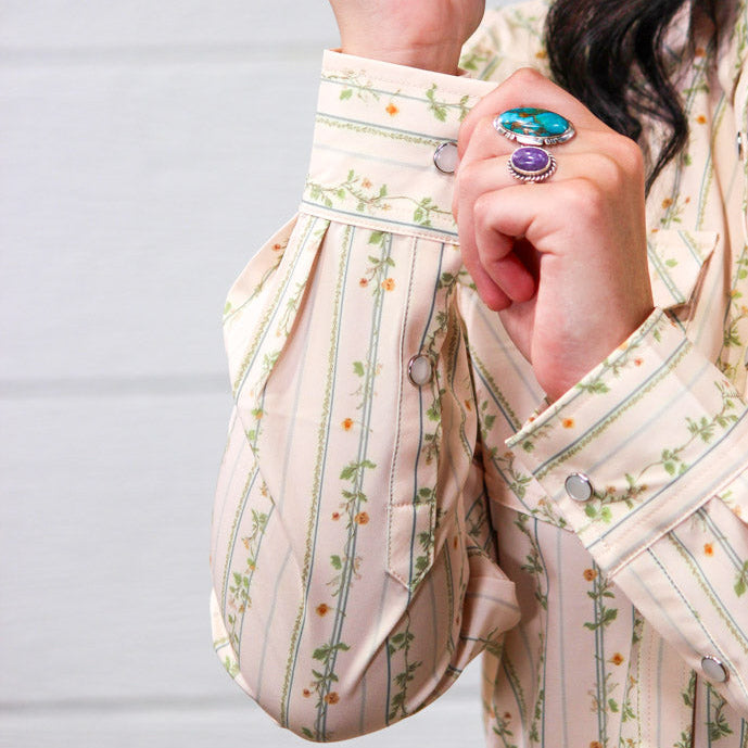 A close-up of a person wearing a cream-colored long-sleeve blouse with vertical green floral stripes and orange flower accents. The person is shown from the chest down, adjusting the cuff of the sleeve, with two rings visible on their fingers against a light gray paneled background.