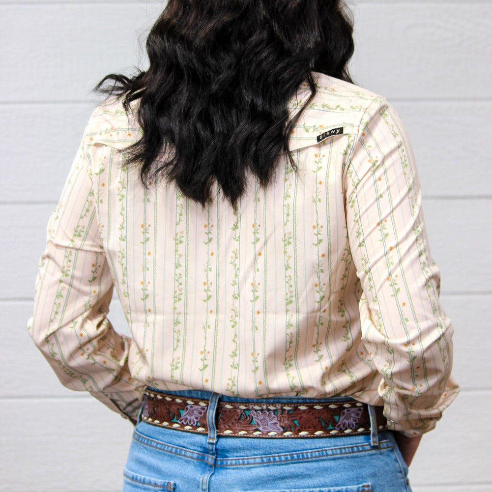 A woman shown from behind wearing a cream-colored long-sleeve western shirt with vertical green and orange floral stripes, paired with a brown embroidered belt and blue jeans, set against a white wood-paneled wall.