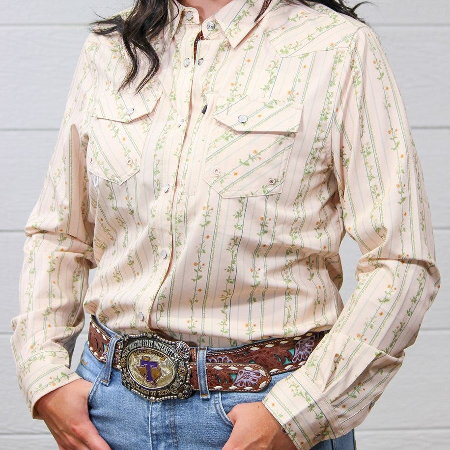 A woman wearing a long-sleeve western-style button-up shirt with vertical floral stripes in beige, green, and orange, paired with a brown embroidered belt and blue jeans against a white wood plank background.