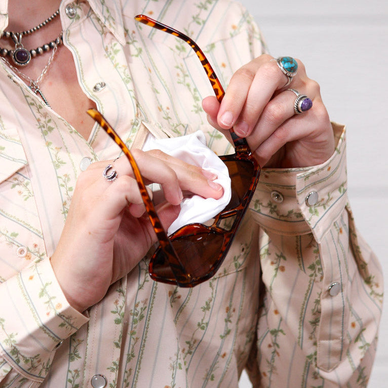 A close-up of a person wearing a cream floral button-up shirt and holding tortoiseshell sunglasses with a white cloth.