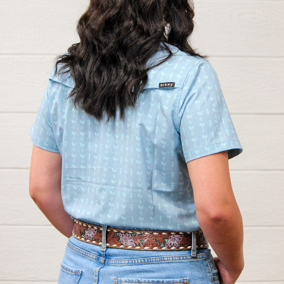 Rear view of a woman wearing a light blue short-sleeve western-style shirt with a small embroidered logo on the back pocket, paired with a brown western belt and jeans against a white paneled wall.