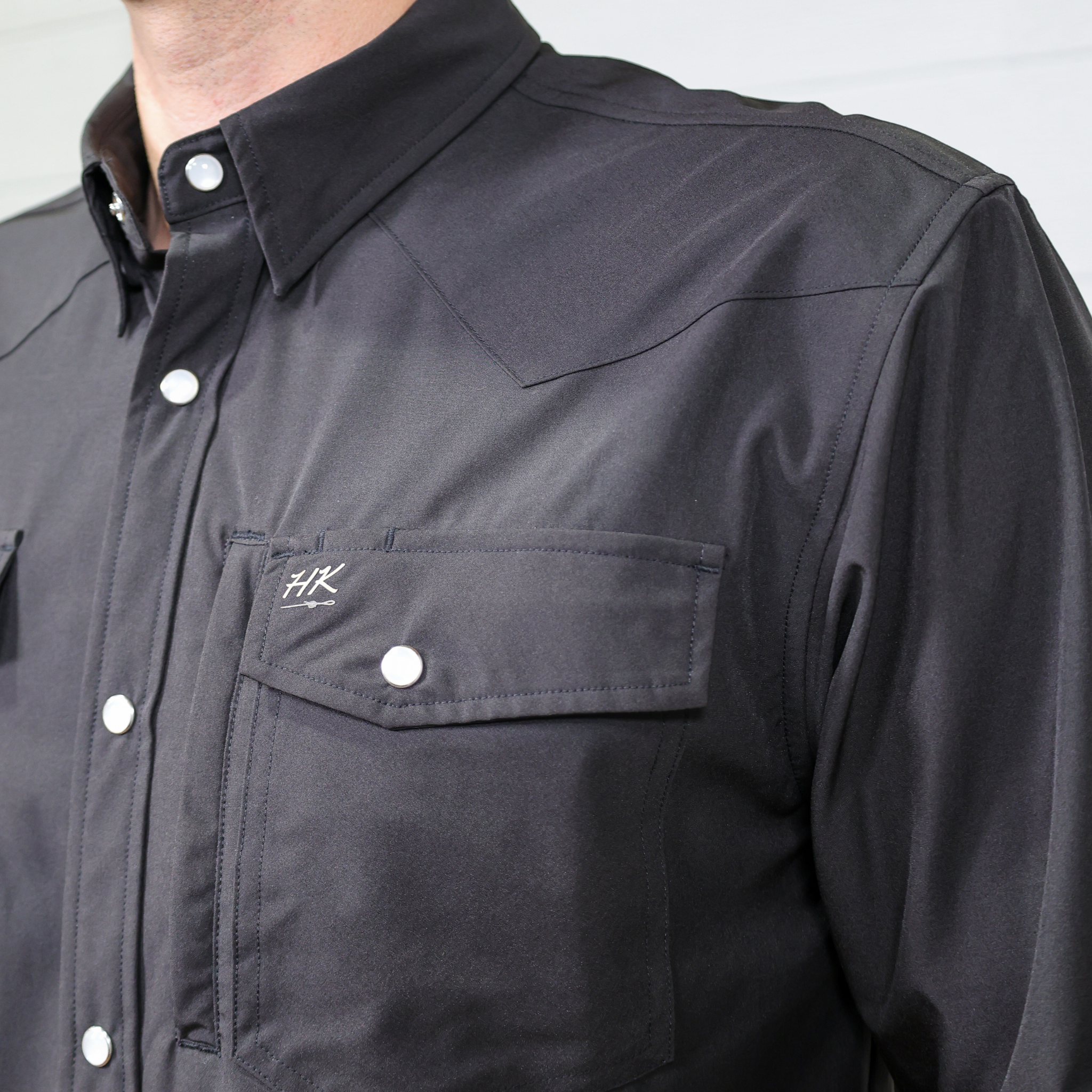 Close-up of a black shirt with a logo on the chest pocket against a white background. Side Facing