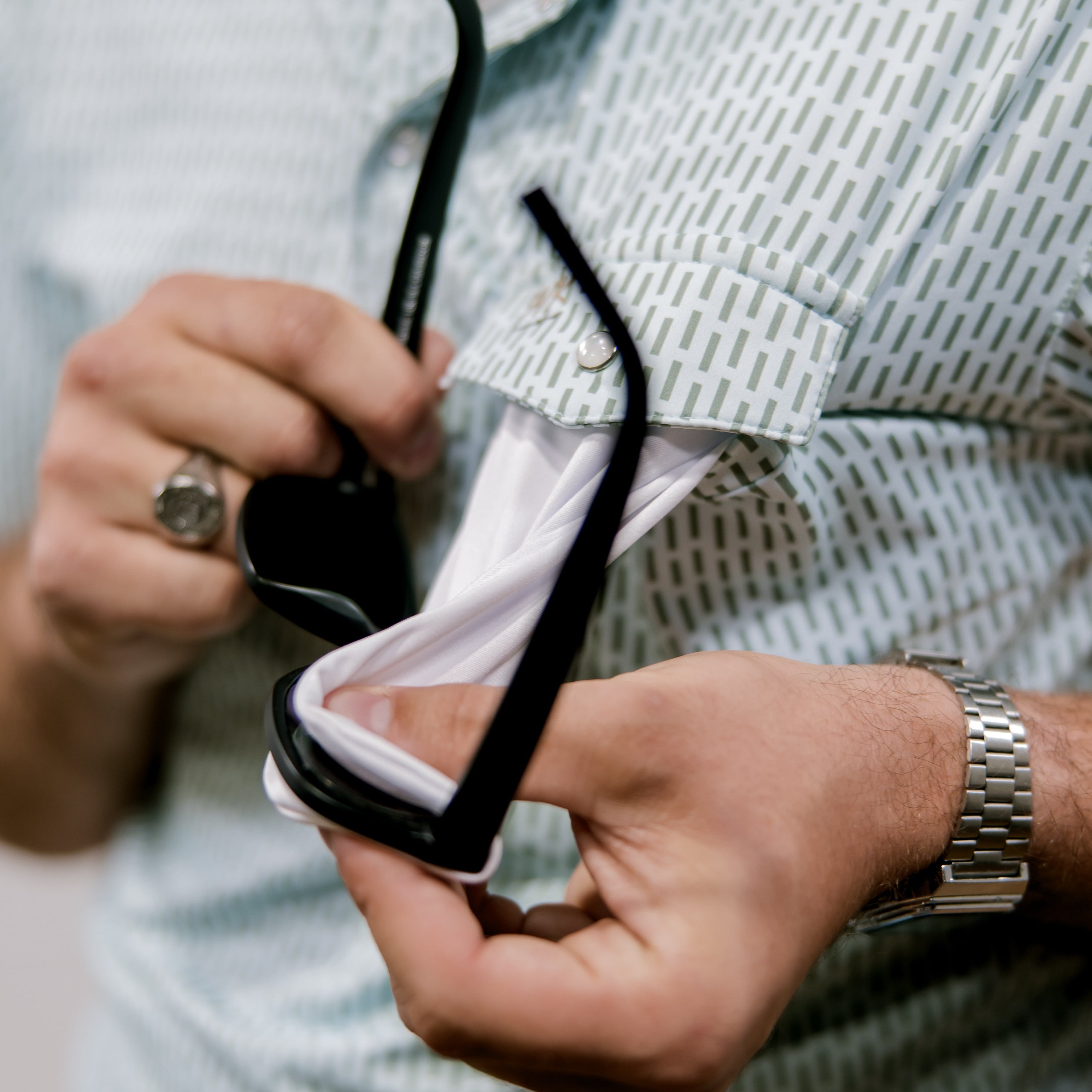 Person cleaning a pair of sunglasses and wearing and blue and green polo with a blurred background.